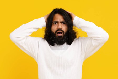 Hombre indio de pelo largo y barba se levanta sobre un vivo telón de fondo amarillo, su expresión de intensa frustración, tocándose la cabeza