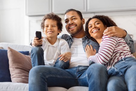 Téléchargez les photos : Une famille afro-américaine de trois personnes, dont deux parents et un enfant, est assise sur un canapé dans un salon. Ils tiennent une télécommande, probablement en regardant la télévision ensemble. - en image libre de droit