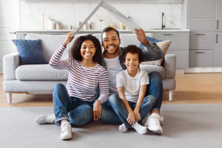 A joyful African American family consisting of mother, father, and their young son is comfortably seated on the floor of their well-lit living room, holding paper cutout of house above their heads