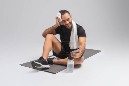 A man is seated on a floor mat with a towel draped around his neck. He appears relaxed and may have just finished exercising or working out, checking smartphone
