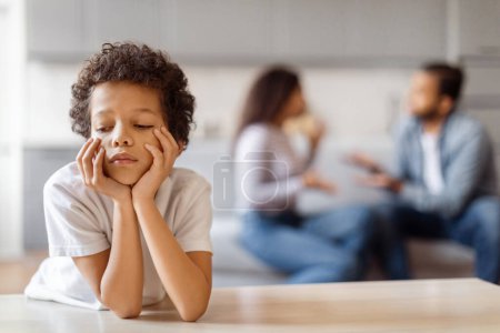 Un jeune enfant afro-américain est assis à une table, les mains couvrant son visage. Il semble être profondément dans la pensée ou la contemplation tandis que ses parents se disputent