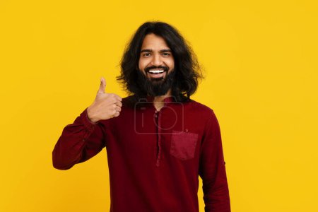 Photo for Indian man with long hair and a beard is enthusiastically giving a thumb up gesture, isolated on yellow studio background - Royalty Free Image