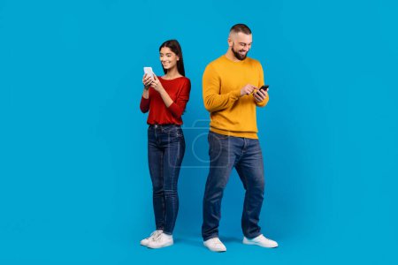 A man in a yellow sweater and jeans and a woman in a casual red top with dark denim jeans stand side by side, each absorbed in their own smartphone, blue background