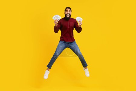 Indian man wearing a red sweater and blue jeans exuberates happiness while jumping in the air. He is holding a bunch of money in both hands, celebrating probably some sort of financial success