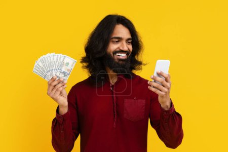 Un Indien aux cheveux longs et à la barbe avec de l'argent et un portable dans les mains. Il semble s'engager dans une transaction ou une communication. Son expression est concentrée et attentive.