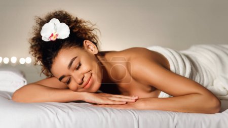 Photo for A serene image featuring an african american woman lying down with a white flower in her hair, looking peaceful in a spa setting - Royalty Free Image