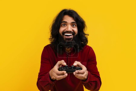Indian man with long hair and a beard holding a video game controller, focusing intently on the screen as he plays a video game, yellow background