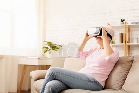 Téléchargez les photos : La femme âgée est assise confortablement sur un canapé beige dans une pièce lumineuse et aérée, avec un casque de réalité virtuelle sur la tête. Elle porte des vêtements décontractés et semble engagée dans le contenu VR - en image libre de droit