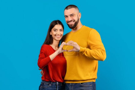 A cheerful man and woman stand close together against a vivid blue backdrop, their fingers intertwined to form a heart shape, symbolizing love and connection