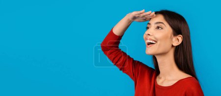 Téléchargez les photos : Une jeune femme joyeuse portant un haut rouge vif se tient debout sur une toile de fond bleue solide. Elle a sa main au-dessus de ses yeux comme si elle les ombrageait du soleil, regardant au loin, copiant l'espace - en image libre de droit