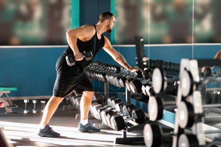 Ein Mann in sportlicher Kleidung ist zu sehen, wie er in einem Fitnessstudio mit einem Hantelständer trainiert. Er stemmt Gewichte mit konzentrierter Zielstrebigkeit und präsentiert Krafttraining als zentralen Bestandteil seiner Fitness-Routine..