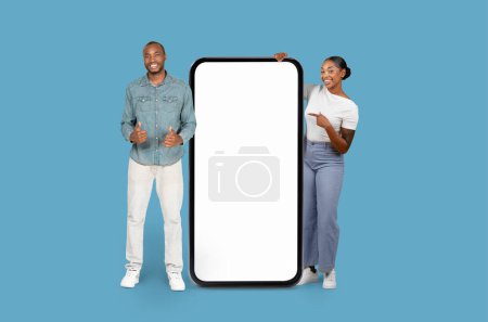 Cheerful African American pair stands by a large smartphone screen for mockups on light blue background