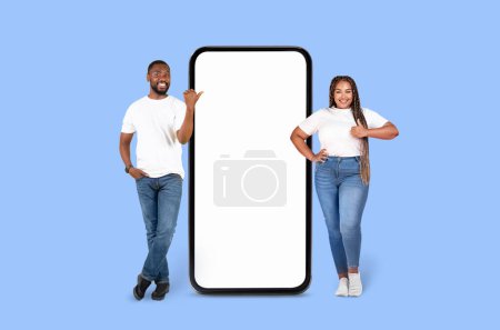 Téléchargez les photos : Heureuse paire afro-américaine posant par une grande maquette d'affichage de téléphone portable sur fond bleu, idéal pour les annonces - en image libre de droit