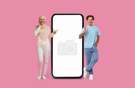 A woman in hijab and man point at blank smartphone screen on a pink background, mockup copy space, mobile app