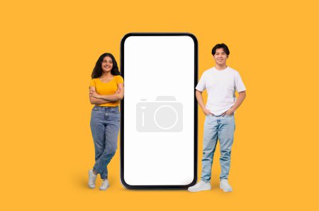 Foto de Hombre joven multirracial y una mujer de pie junto a un gran espacio de copia maqueta smartphone sobre un fondo amarillo - Imagen libre de derechos