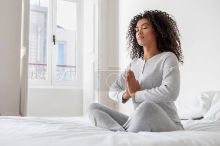 A young Hispanic woman is peacefully meditating with her eyes closed and hands in a prayer position, seated on a white bed in a brightly lit bedroom with natural daylight streaming through the window.