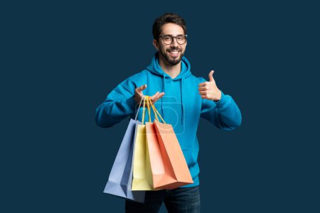 Photo for A man wearing a blue hoodie is standing while holding shopping bags in his hands. He is smiling and giving a thumbs up gesture, expressing satisfaction with his purchases. - Royalty Free Image