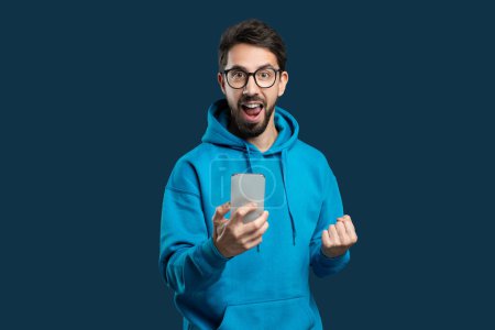 Téléchargez les photos : Un homme portant un sweat à capuche bleu est debout à l'extérieur, tenant un téléphone portable dans sa main. Il semble regarder l'écran attentivement, peut-être en envoyant des SMS ou en naviguant, en faisant des gestes - en image libre de droit