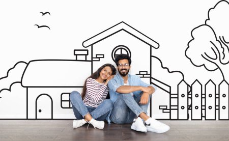 Photo for Cheerful young indian couple sitting by white wall with a house outline, dreaming of a home. Real estate, mortgage concept - Royalty Free Image