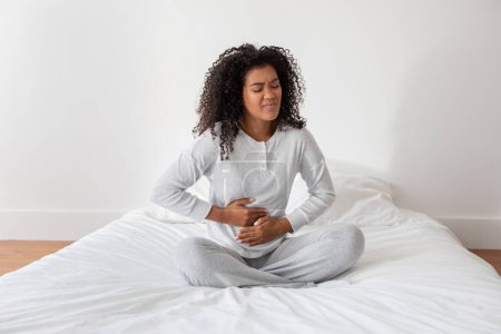 A young Hispanic woman with curly hair sits cross-legged on a bed, clutching her stomach with a pained expression on her face suggesting discomfort or illness