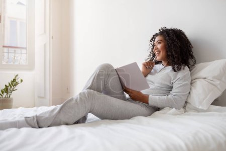 La mujer hispana está sentada en una cama, absorta en la lectura de un libro. Ella sostiene el libro en sus manos, con una expresión enfocada en su cara