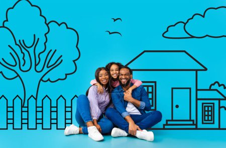 Téléchargez les photos : Une joyeuse famille afro-américaine de trois personnes, avec une mère, un père et un enfant, est assise contre une illustration ludique d'une maison et d'arbres dessinés sur un fond bleu vif - en image libre de droit