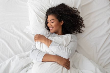 Photo for Hispanic woman with curly hair smiles contentedly as she embraces a fluffy pillow, lying in comfort on a bright white bedspread - Royalty Free Image