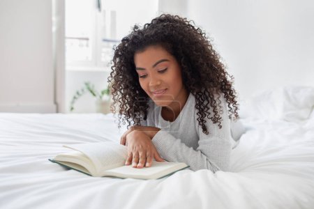 A Hispanic woman is laying comfortably on a bed, engrossed in reading a book. The room is softly lit, and she is fully focused on the text, turning pages occasionally.