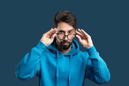 A focused man with a beard and tousled hair is adjusting his glasses and looking straight ahead. He is wearing a vibrant blue hoodie, which contrasts with the solid blue backdrop