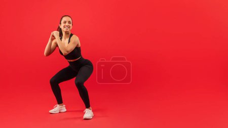 A joyful woman wearing sportswear, including a black tank top and leggings, performs a squat exercise with her hands together, red background, copy space