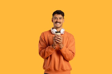 A cheerful man wearing a casual orange sweatshirt stands against a vivid yellow backdrop, holding a white cup in his hands with wireless headphones around his neck