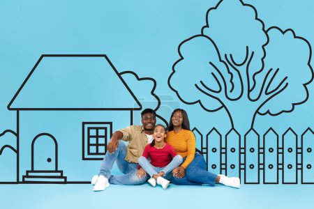 Téléchargez les photos : Une joyeuse famille noire composée de deux adultes et d'un enfant sont assis étroitement les bras l'un autour de l'autre devant une illustration dessinée d'une maison et d'un arbre sur un fond bleu uni - en image libre de droit