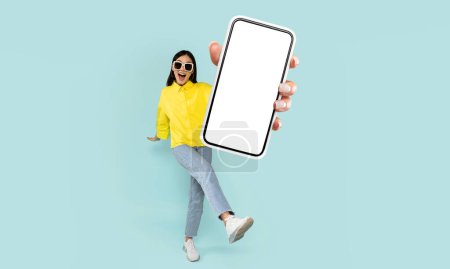 Photo for A joyful woman wearing sunglasses and a bright yellow shirt is leaning towards the camera, showcasing a smartphone with a blank screen, ready for content placement. - Royalty Free Image