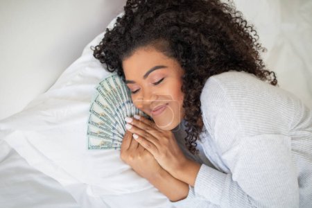 A Hispanic woman is seen laying on a bed with a large amount of money spread out around her. She appears to be counting or looking at the money.