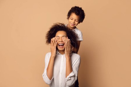 African American young child is gleefully covering his mothers eyes from behind as they both share a moment of joy and surprise