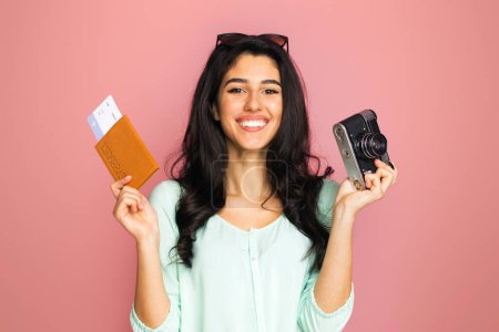 Photo for A middle eastern woman is standing while holding a camera and a passport. She appears to be ready for traveling or documenting her journey - Royalty Free Image