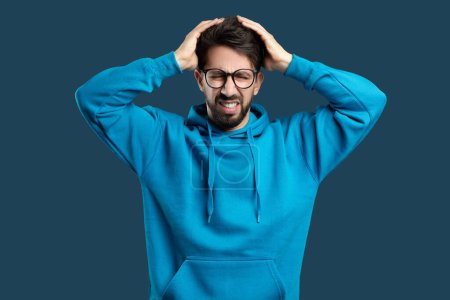 A young man with a beard and glasses, wearing a blue hoodie, is standing against a solid blue background. He appears frustrated or stressed