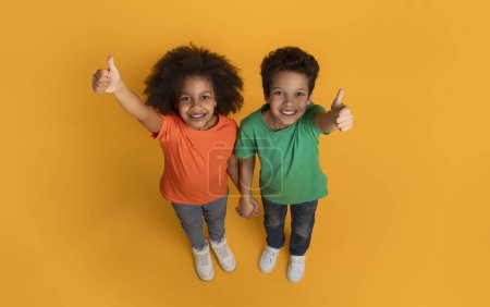 A cheerful African American boy and girl are standing on a bright yellow surface, looking upwards into the camera with joyful smiles, giving a thumbs up gesture, top view
