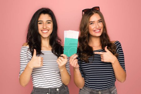 Zwei fröhliche junge Frauen, eine mit dunklem lockigem Haar und die andere mit welligem braunem Haar, halten Reisepass und Flugtickets in der Hand und zeigen mit erhobenen Daumen darauf.
