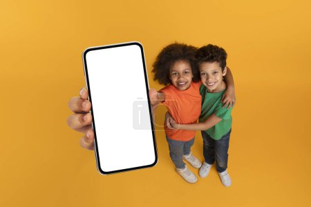 Foto de Niños afroamericanos sosteniendo un teléfono celular con una pantalla blanca en blanco, que no muestra contenido o imágenes, espacio de copia de maqueta, fondo amarillo del estudio - Imagen libre de derechos
