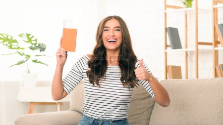Photo for A delighted young woman stands in a sunny living room, waving her passport and giving a thumbs up, expressing her eagerness to embark on a journey - Royalty Free Image