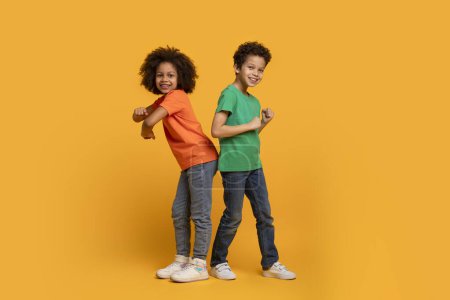 A pair of joyful African American children, a boy and a girl, are standing back to back with confident smiles on their faces, dancing on yellow background