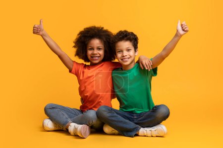 Two African American children are sitting on the floor with their arms stretched up in the air, showing thumb ups. They look joyful and energetic as they play together.