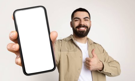 Millennial man is holding a smart phone with white blank screen in his hand, showing thumb up, isolated on beige studio background