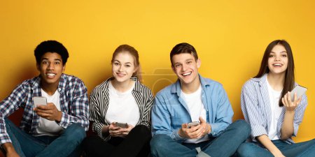 Photo for A cheerful quartet of young friends are seated side by side, casually dressed, each holding a smartphone, amused and engaged in a moment of digital interaction, against a vibrant yellow backdrop - Royalty Free Image
