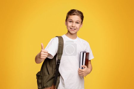 Foto de Buena educación doméstica. Alegre adolescente chico con mochila y libros gesto pulgar hacia arriba, fondo de estudio naranja - Imagen libre de derechos