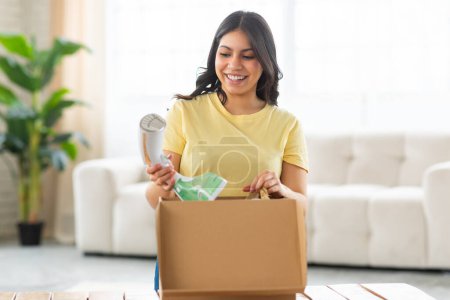 Photo for A cheerful middle eastern woman is standing in a brightly lit living room, looking delighted as she unpacks modern iron from a cardboard box. - Royalty Free Image