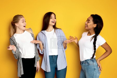Three multiethnic girls are standing side by side with one of them gesturing animatedly as if in the middle of an engaging story, yellow studio background