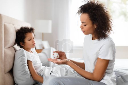 A concerned African American mother is gently offering medication to her unwell young son, who is resting on a bed in a sunlit room