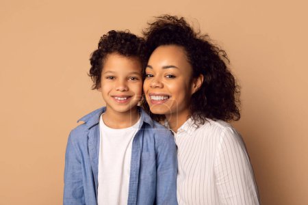 African American woman and a child are standing together, smiling and posing for a camera. The woman has her arm around the child, both looking directly at the camera.
